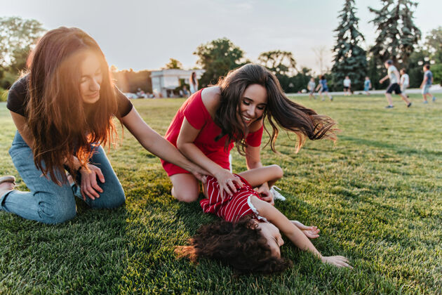 草地两个黑发女孩坐在草坪上和她们的小妹妹玩耍周末女士们和孩子们在一起的户外照片放松公园拥抱