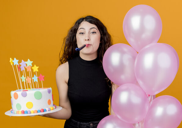 哨子年轻漂亮的女人卷发拿着生日蛋糕和一堆气球吹口哨快乐和兴奋的生日派对概念站在橙色的墙上吹卷发兴奋