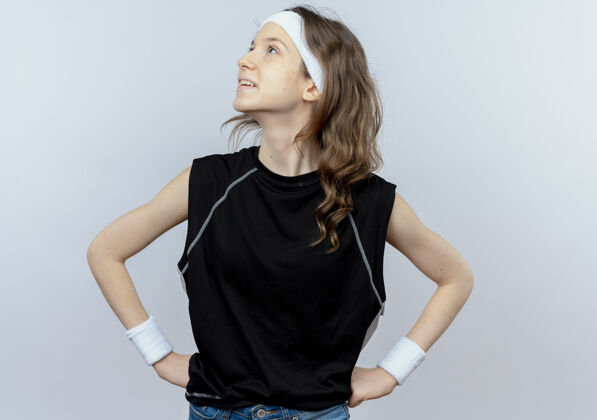 运动装身穿黑色运动服的年轻健身女孩戴着头巾 面带微笑 手臂放在臀部 站在白色的墙上微笑年轻靠边站