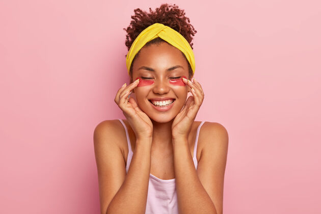 眼睛年轻的非洲裔女模特在眼睛下面涂上胶原蛋白护垫 享受保湿治疗 笑容灿烂 露出洁白的牙齿 有新鲜健康的皮肤 戴着黄色的头巾恢复水凝胶健康