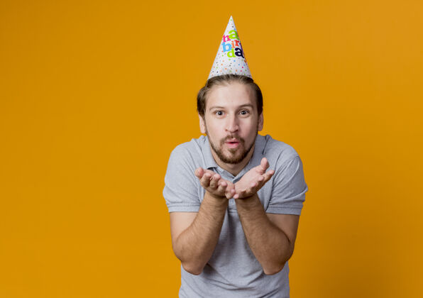 吻戴着节日帽的年轻人庆祝生日聚会快乐而积极地站在橙色的墙上 在他面前用手吹着吻年轻帽子前面