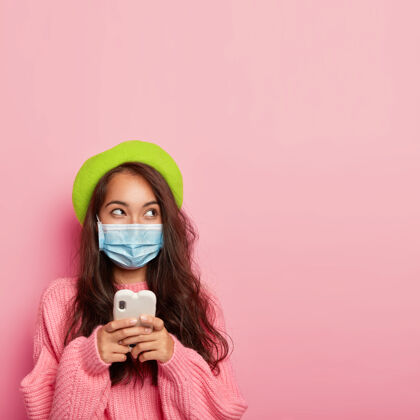 疾病千禧一代的亚洲女人拿着手机站着 想些什么 往一边看 保护自己不受季节性病毒感染 穿着粉色针织毛衣在室内摆姿势沉思防护女性