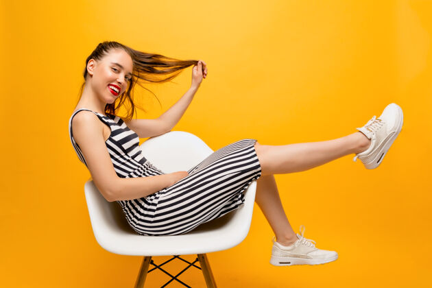 肖像时尚快乐的红唇女人的完整长度照片 头发穿着合身的裙子和白色的运动鞋坐在椅子上 玩她的头发在黄色的墙壁上 文本的地方休闲欢呼女孩