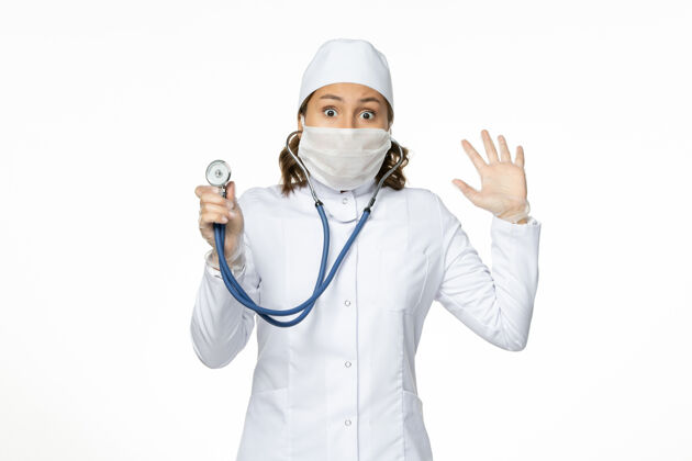 正面正面图女医生穿着白色医疗服 戴着口罩 在白墙上用听诊器给大流行病毒治病护士病毒制服