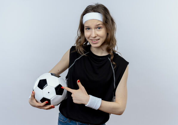 运动装身穿黑色运动服的年轻健身女孩 头戴钢带 手拿足球 手指指向足球 站在白色的墙上开心地微笑着抱着手指足球