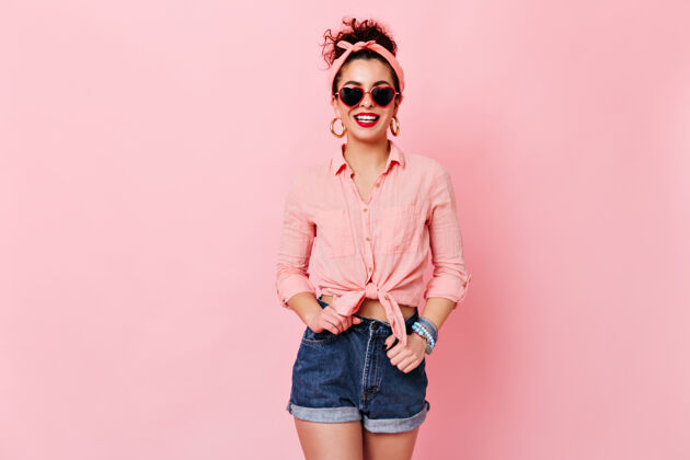 调情心情很好的女人在粉红色的空间里摆姿势戴着墨镜和金耳环 穿着衬衫和短裤的女孩在微笑发型太阳镜DIVA
