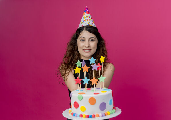 积极年轻漂亮的女人 卷发 戴着节日帽 手里拿着生日蛋糕 微笑着 站在粉色的墙上女人年轻站着