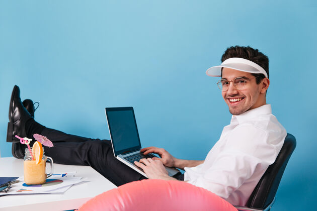 帽子一个穿着白衬衫 戴着帽子和眼镜的迷人男人正对着蓝色的空间那个家伙拿着笔记本电脑 在度假时工作成功正式工作