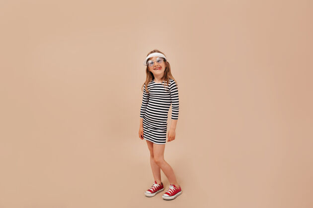 孩子全长度的照片可爱迷人的小女孩穿着裙子 帽子和运动鞋摆在相机在米色背景微笑幼儿全长