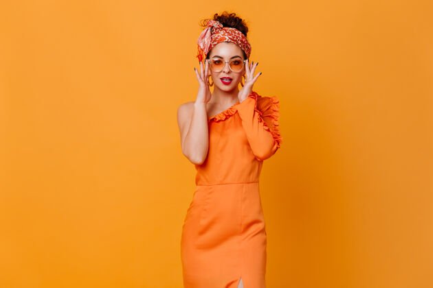 女士时尚的黑发女士戴着非洲风格的头带 戴着墨镜 穿着优雅的裙子 在橙色的空间里风骚地看着镜头橙色连衣裙现代卷曲