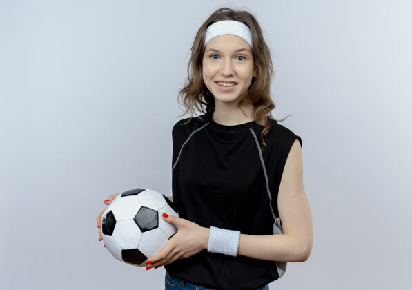健身身穿黑色运动服的年轻健身女孩戴着头巾 面带微笑地举着足球站在白色的墙上年轻抱头带
