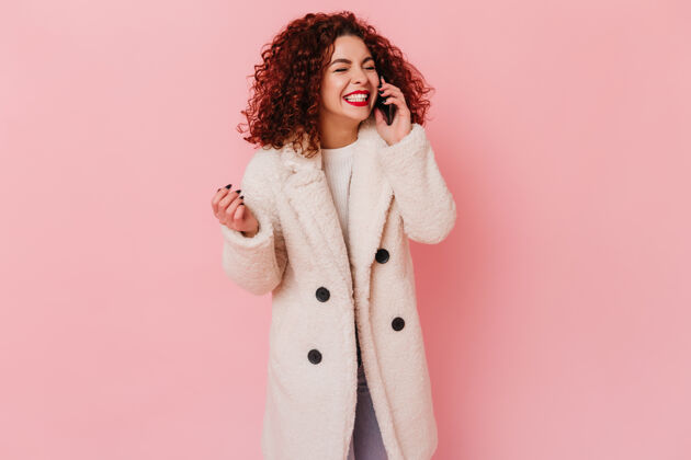 魅力快乐的卷发黑发女孩穿着白色环保外套在粉红色的空间愉快地讲电话时尚优雅漂亮
