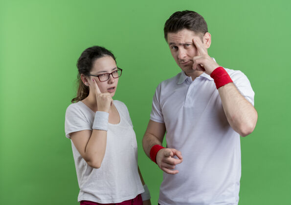 手指一对年轻的运动情侣手指着太阳穴专注于一项艰巨的任务站在绿色的墙壁上女人夫妇立场