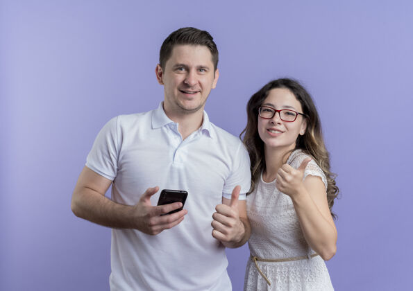 男人一对年轻夫妇站在蓝色的墙上 看着手持智能手机的照相机 竖起大拇指女人抱着手机