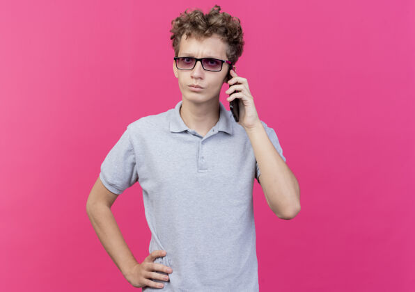 穿一个戴着黑眼镜 穿着灰色马球衫的年轻人站在粉红色的墙上 严肃地讲着手机眼镜年轻马球