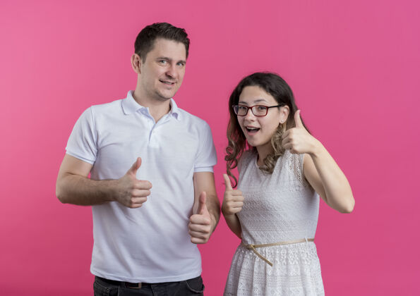 站立一对穿着休闲服的年轻夫妇站在粉红色的墙上 兴高采烈地笑着竖起大拇指表演微笑男人