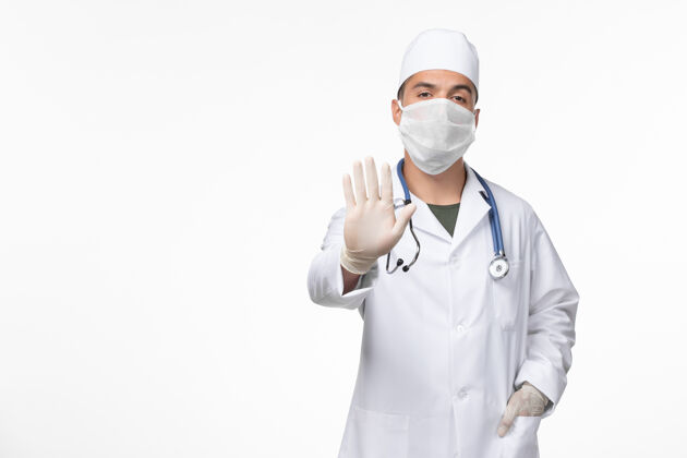 病毒正面图：男医生穿着医疗服 戴着口罩 戴着听诊器 在浅白的办公桌上对着病毒-病毒-疾病大流行实验室外套面罩视图