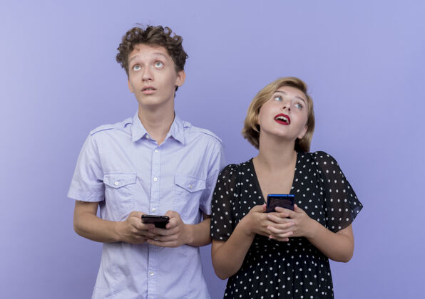 移动一对年轻漂亮的夫妇拿着智能手机站在一起 在蓝色的墙上显得很困惑抱着站着夫妻