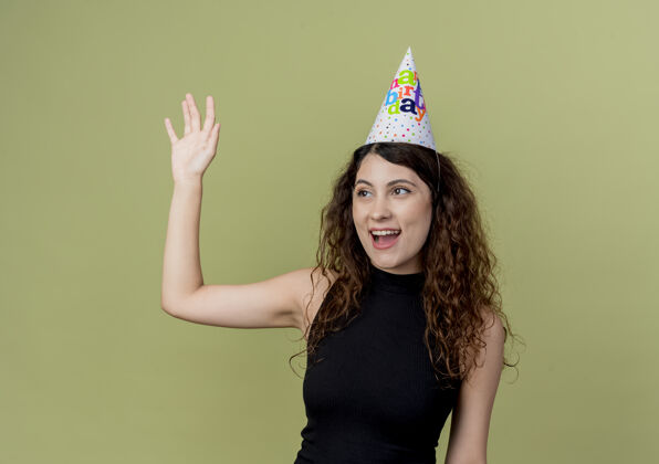光年轻漂亮的女士 一头卷发 戴着节日帽 快乐而积极地挥手 生日聚会的概念站在光墙上手女人帽子