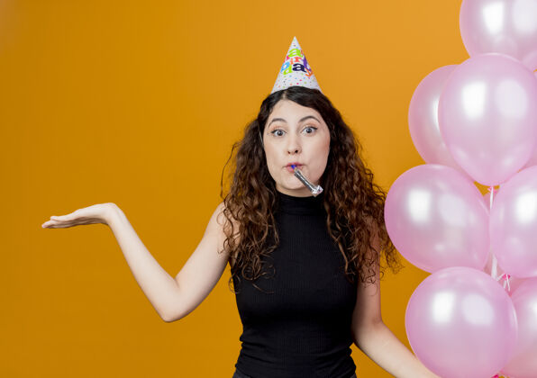 帽子一个年轻漂亮的女人 卷发 戴着节日帽 手里拿着气球吹着口哨 看起来像是在交谈 手臂伸向一边 站在橙色的墙上 生日派对的概念年轻吹站