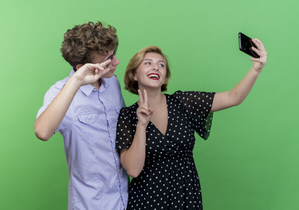 使用年轻漂亮的夫妇站在一起用手机自拍微笑着在绿色的墙上展示v标志展示采取Vsign