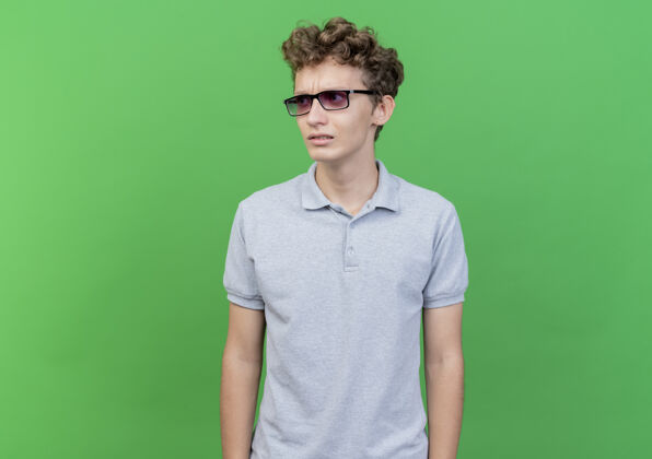 男人一个戴着黑眼镜 穿着灰色马球衫的年轻人站在绿色的墙边 心神不定地站在一边靠边站担心年轻