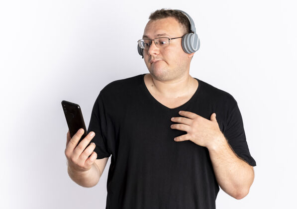 耳机一个戴眼镜的超重男人 穿着黑色t恤 戴着耳机 拿着智能手机 站在白色的墙上 困惑地看着屏幕困惑屏幕手机