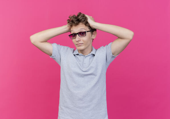 失望一个戴着黑眼镜 穿着灰色马球衫的失望的年轻人 站在粉红色的墙上摸着他的头 感到困惑站着年轻马球