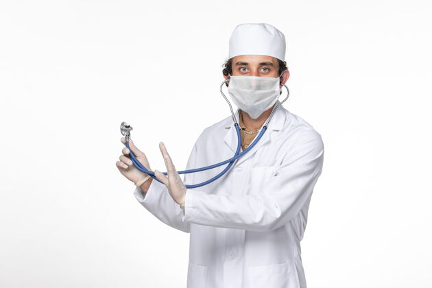 套装正面图：男医生穿着医疗服 戴着口罩 以防感染冠状病毒-在白色桌面上使用听诊器飞溅冠状病毒大流行专业医生视图
