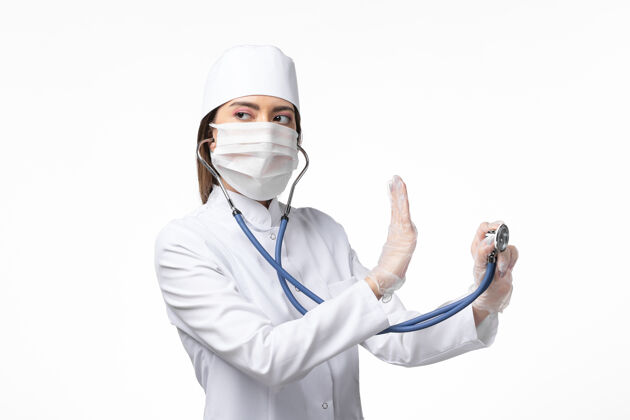 专业人士正面图女医生穿着白色医疗服 戴着口罩 因大流行手持听诊器 白色桌面上有疾病药物病毒大流行病毒covid-实验室外套正面听诊器