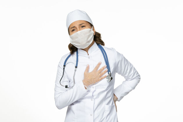 医疗白壁上有心痛的身穿白色医疗服和口罩的女医生pandemy病毒病药专业听诊器疾病