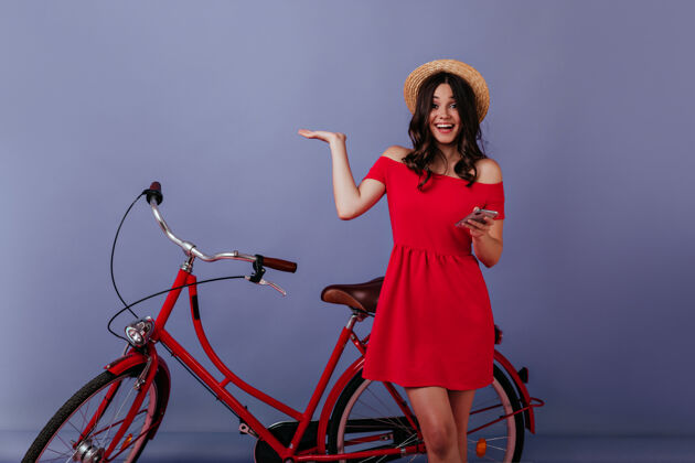 棕色头发兴奋的女人手里拿着电话站在自行车旁边情绪激动的黑发女孩戴着草帽在自行车前摆姿势快乐休闲活动自行车