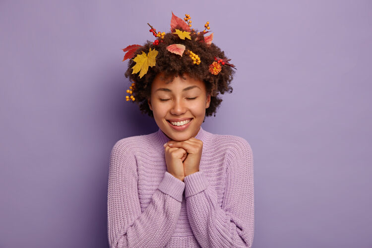 紫色可爱快乐女人的室内画像双手放在下巴下 享受温暖干燥的秋日 感受快乐 头发里有树叶和罗汉浆果 穿着毛衣 隔离在紫色的墙上秋天秋天表情