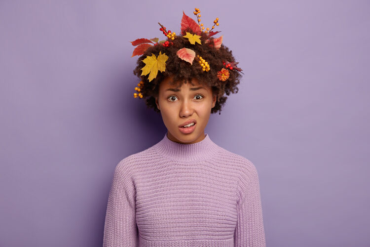 忧郁可爱的不满的非裔美国女士微笑着面对 钱包嘴唇 有悲伤的脸表情 穿着黄色的叶子和浆果在头发针织毛衣孤立的紫色美国秋天紫色