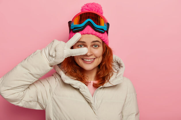 帽子微笑的女性 戴着冬天的帽子和夹克 戴着白色的手套 在眼睛上做着和平的手势 有季节性的休息 隔离在粉红色的墙上护目镜室内红发