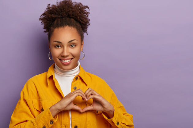 温柔善良可爱的非裔美国妇女做心脏手势 承认爱情 表达同情和喜悦 有真实的感情 穿着时尚的衣服 孤立在紫色的背景下同情标志象征
