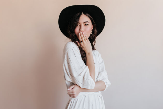 服装戴着黑帽子的沉思的亚洲女人看着相机在米色背景上孤立的迷人的日本模特的摄影棚拍摄配饰爱韩语