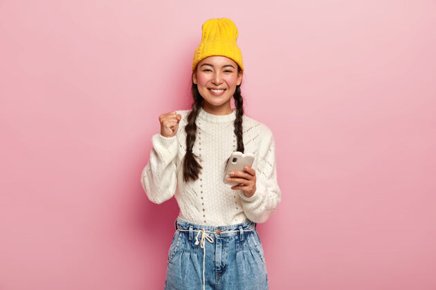 手机开朗的韩国年轻女子握紧拳头 戴着黄色时髦的帽子和白色的毛衣 愉快地微笑着 手持现代手机 隔离在玫瑰色的工作室墙上微笑拳头室内