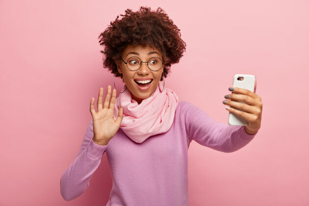 黑发乐观微笑的女人打招呼记录视频信息 通过手机上的社交媒体应用程序打电话 手持小玩意在前面 挥动手掌 穿着时髦的服装围巾表情眼镜