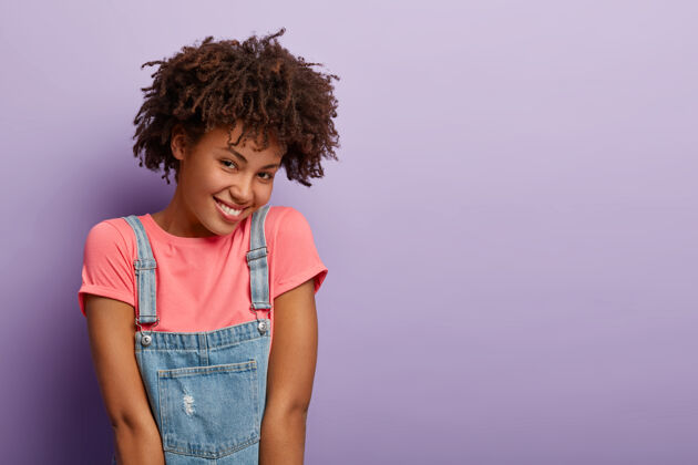 非洲快乐的非洲裔美国女性有着开朗害羞的表情 表达积极的情绪 穿着粉色t恤和牛仔裤 模特在紫色的墙上 复制空间在一旁牙齿休闲时尚