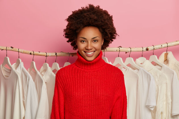 购物快乐的黑皮肤女造型师 非洲式发型 笑容灿烂 穿着红色针织毛衣 站在衣架旁 带着衣架喜悦套头衫消费者