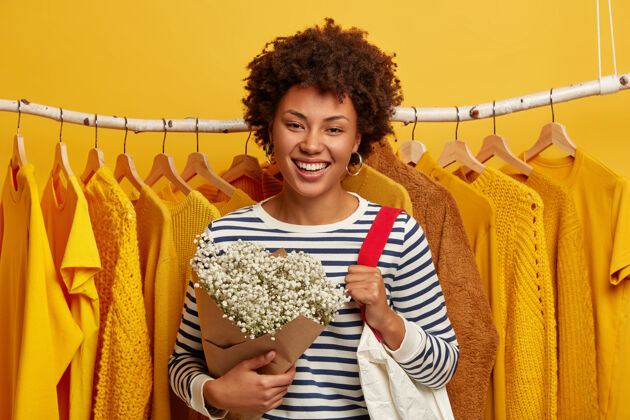 一女人的生活方式 时尚 消费主义的概念美国黑人女人脸上带着喜悦的表情 站在服装店 肩上扛着购物袋 从丈夫那里得到鲜花 黄色背景购买衣架高兴
