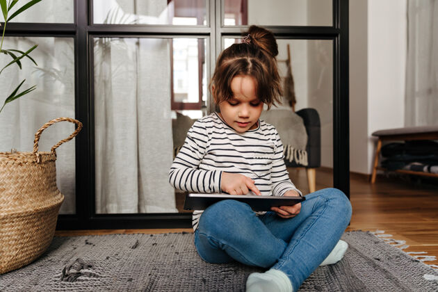 观看穿着紧身牛仔裤和条纹毛衣的小女人在平板电脑里画画 坐在客厅的地板上编辑在线学习无线