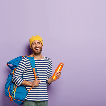 背包快乐无胡须男背包客的垂直镜头背着大旅游背包 拿着烧瓶 隔着紫罗兰色的墙 自由的空间人和旅游白种人一个服装