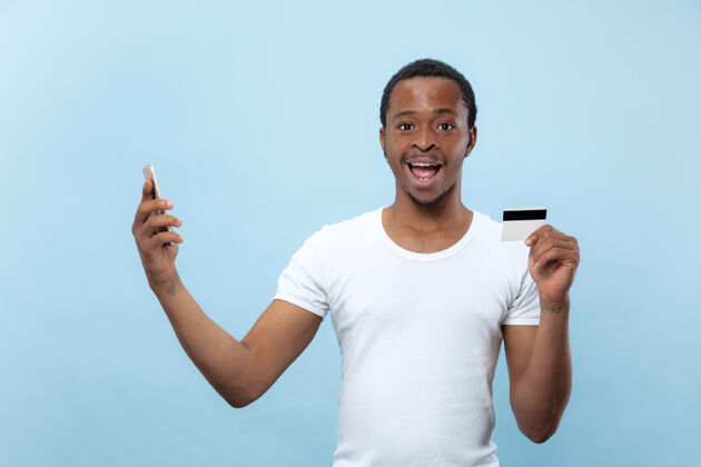 企业家在蓝色空间里 一个身穿白衬衫 手持卡片和智能手机的年轻非裔美国人的半身肖像支付订单面部