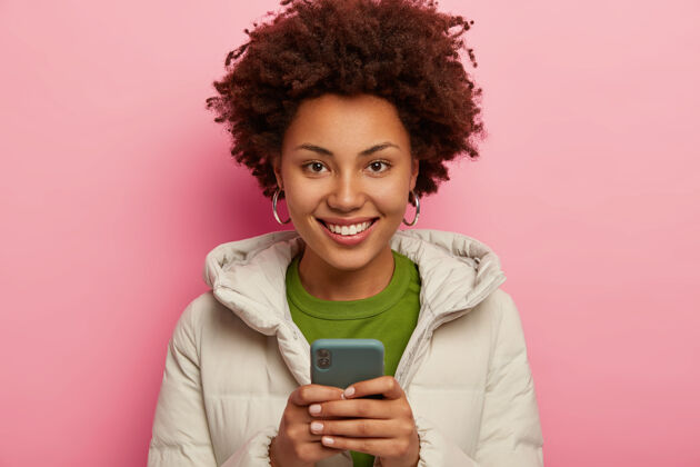 短信喜黑皮肤的潮人女孩画像用手机 查看邮箱 穿着暖和的冬衣 隔着粉色的墙壁 社交网站冬天使用举行