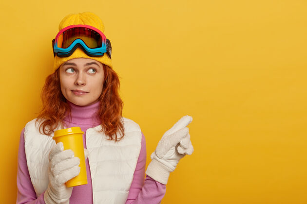 饮料姜黄色头发的滑雪者 戴着黄色帽子 指着旁边的空地 拿着外卖咖啡 展示冬天的景色 站在黄色的背景下感冒运动型指尖