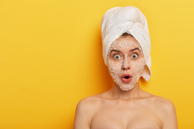 沐浴尴尬的年轻女子站在赤裸的脸上 用去皮的面膜去掉脸上的暗宠 盯着相机上戴着白色软毛巾的头上的黄色卫生印象面膜