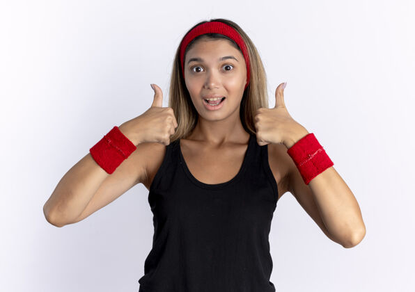 站身穿黑色运动服 头戴红色头巾 满脸幸福 竖起大拇指的年轻健身女孩站在白墙上女孩拇指年轻