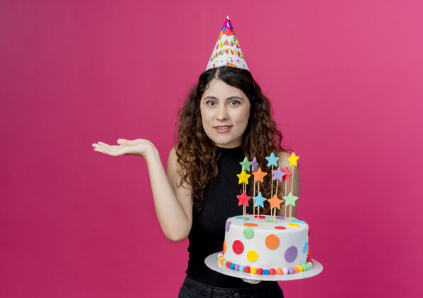 困惑年轻漂亮的女人卷发戴着节日帽拿着生日蛋糕困惑地耸耸肩站在粉色的墙上生日派对的概念女人年轻看着